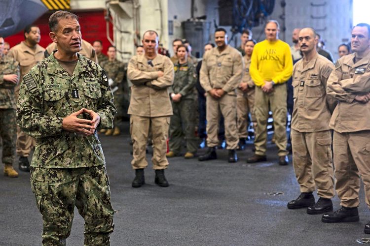 Ein Offizier der U.S. Navy in einer Tarnuniform spricht vor einer Gruppe von Besatzungsmitgliedern auf einem Schiff. Die Anwesenden hören aufmerksam zu, teils in beigen Uniformen, teils in ziviler Kleidung. Der Hintergrund zeigt das Innere eines Schiffs.