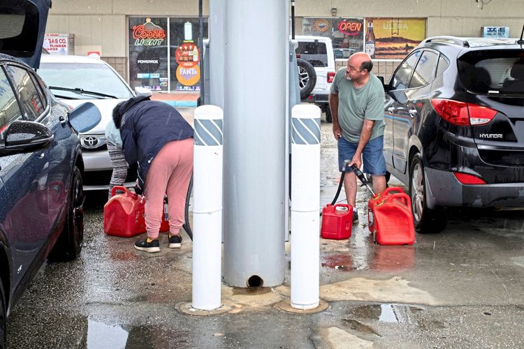 Zwei Personen füllen rote Benzinkanister an einer Tankstelle auf. Um sie herum stehen Autos geparkt, und im Hintergrund sind Geschäfte mit leuchtenden Schildern zu sehen. Es ist regnerisch, und der Boden ist nass.