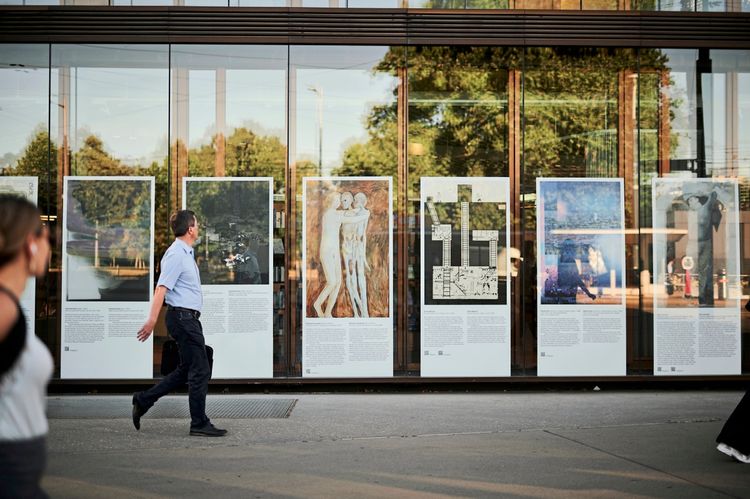 Eine Reihe von Informations- und Kunsttafeln ist vor der Glasfassade eines Gebäudes aufgestellt. Darauf befinden sich Texte und Bilder verschiedener Künstler:innen. Im Vordergrund gehen Menschen vorbei, darunter ein Mann, der von links nach rechts läuft. Im Glas spiegeln sich Bäume und die Umgebung.