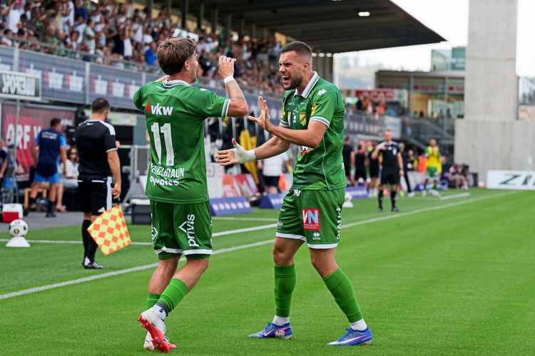 Zwei Fußballspieler des SC Austria Lustenau jubeln über ein Tor während eines Bundesliga-Spiels gegen RZ Pellets WAC in Lustenau. Der Spieler rechts scheint der Torschütze zu sein. Zuschauer, Schiedsrichter und Ersatzspieler sind im Hintergrund zu sehen.