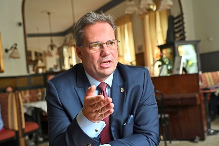 Ein Mann in einem dunklen Anzug mit Brille sitzt in einem Café-ähnlichen Raum und gestikuliert während eines Gesprächs. Im Hintergrund sind Sitzmöbel und Dekorationen erkennbar.