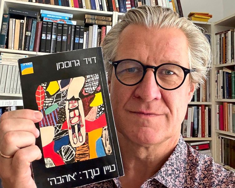Ein Mann mit grauen Haaren, schwarzer Brille und gemustertem Hemd hält ein Buch von David Grossmann mit hebräischem Titel und buntem Cover in der Hand. Im Hintergrund sind Bücherregale mit zahlreichen Büchern zu sehen.