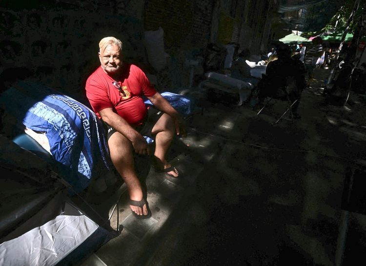 Ein Mann mit blondem Haar sitzt auf einem Campingstuhl im Freien, inmitten von schattigen und sonnendurchfluteten Bereichen. Hinter ihm sind blaue Decken und Umrisse von Zelten oder Campingequipment sichtbar. Die Umgebung deutet auf eine improvisierte Situation hin.
