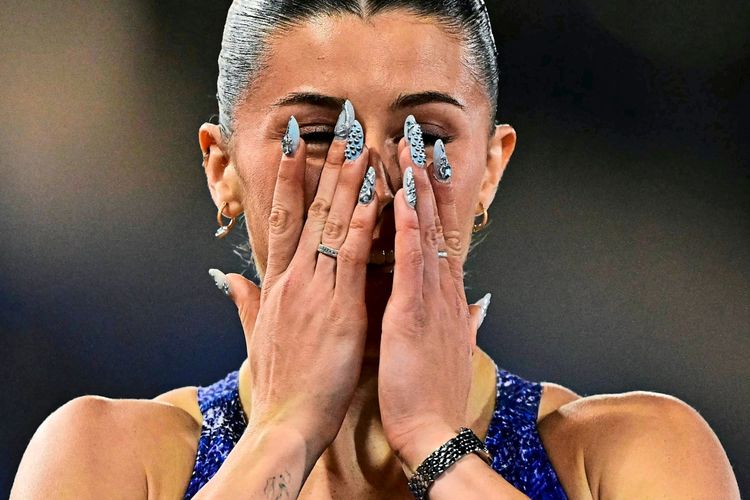 Eine Frau in Sportkleidung hält ihre Hände vor ihr Gesicht, offenbar emotional ergriffen. Sie trägt auffälligen Nagellack und Schmuck. Hintergrund unscharf.
