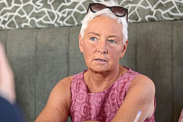 Eine ältere Frau mit kurzem, weißem Haar und Sonnenbrille auf dem Kopf sitzt an einem Tisch. Sie trägt ein ärmelloses, rosa Oberteil und schaut aufmerksam in eine Richtung. Vor ihr liegt ein Notizbuch, in das eine andere Person schreibt. Im Hintergrund ist eine grüne Wand mit abstraktem Muster zu sehen.