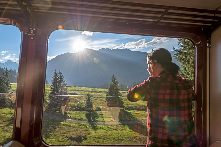 Eine Frau schaut aus dem Fenster im Zug. Sie fährt vorbei an einer idyllischen Landschaft.