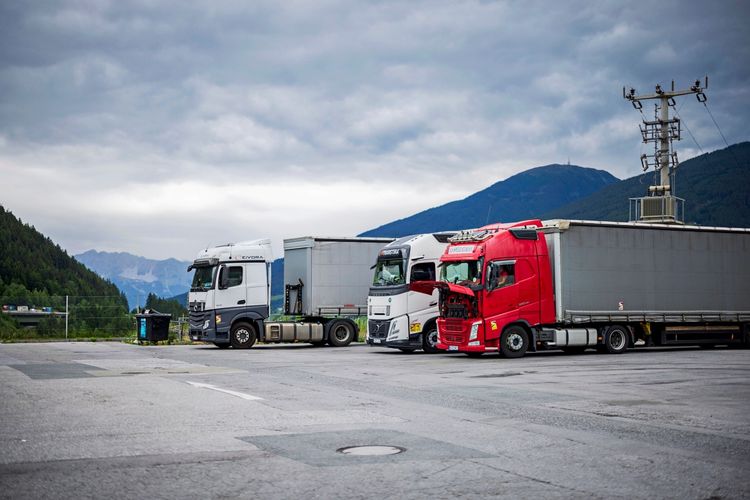Drei Lkw, parkend auf einem Rastplatz am Brenner. Im Vordergrund ist ein roter Lkw mit geöffneter Motorhaube zu sehen, dahinter stehen ein weißer und ein grauer Lkw. Der Hintergrund zeigt bewaldete Berge unter einer wolkigen Himmel.