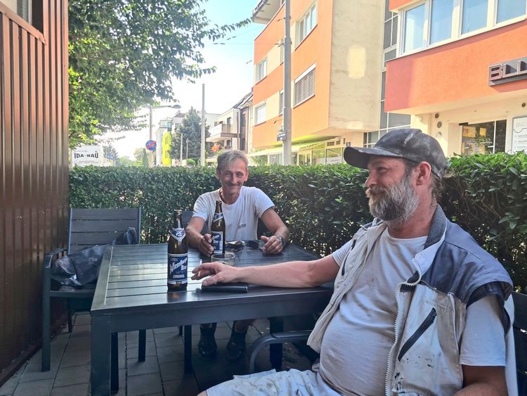 Zwei Männer sitzen an einem Tisch im Freien, umgeben von Grünpflanzen. Beide tragen Freizeitkleidung, einer eine Schirmmütze. Auf dem Tisch stehen Bierflaschen. Im Hintergrund sind Gebäude und eine Straße zu sehen.