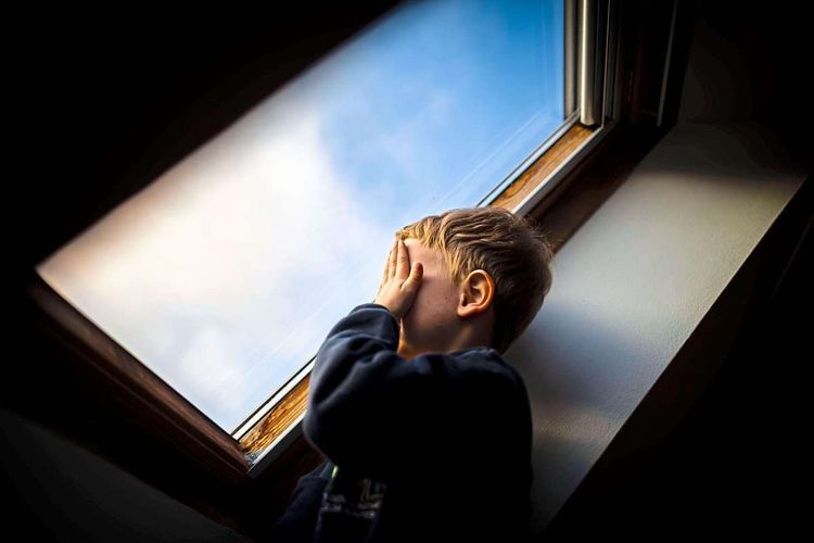 Kind steht am Fenster und verdeckt die Augen mit den Händen. 