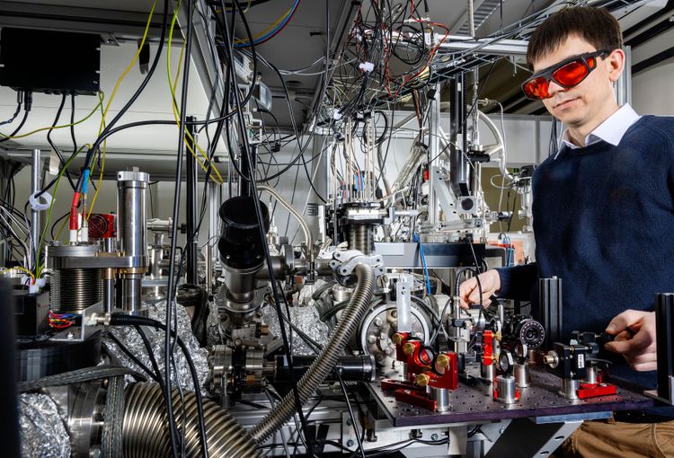 Forscher mit kurzen braunen Haaren trägt Schutzbrille mit roten Gläsern und steht vor einer Anlage mit vielen Kabeln und Röhren.