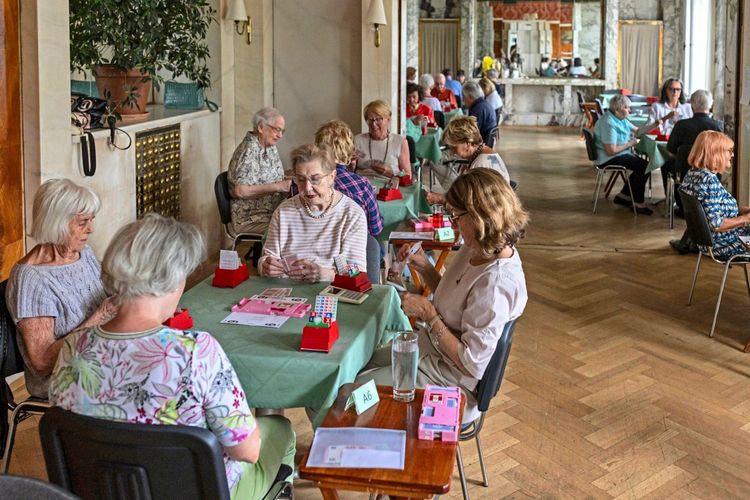Personen unterschiedlichen Alters sitzen in einem eleganten Raum an Tischen und spielen Bridge. Die Szene wirkt entspannt, der Raum ist mit Spiegeln, Pflanzen und Holzboden ausgestattet. Im Vordergrund sieht man Spielkarten und ein Spielgerät auf einem Tisch mit grünem Tischtuch.