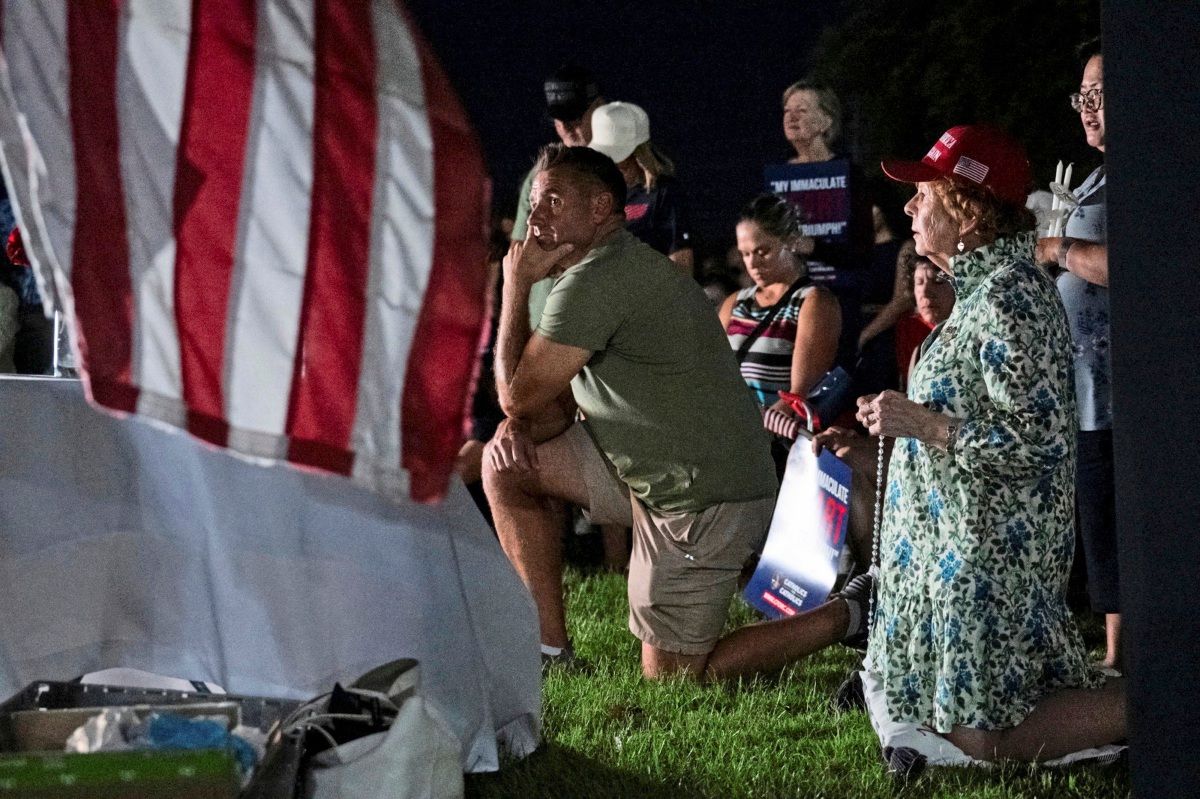 A group of people kneels on a lawn at night, some praying. In the foreground is a section of a US flag. A woman wears a floral dress and red hat, next to her kneels a man in shorts and a t-shirt. In the background, people hold signs.
