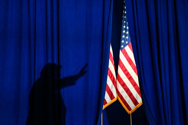 Eine Silhouette von US-Präsident Donald Trump. ist hinter einem blauen Vorhang sichtbar, während rechts die US-Flagge zwischen den Vorhängen steht.