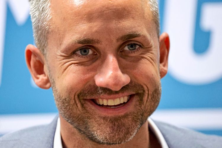 Ein lächelnder Mann mit kurzem, grauem Haar vor einem unscharfen, blau-weißen Hintergrund.