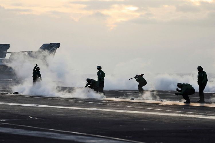 Flugdeckpersonal arbeitet auf dem Deck des Flugzeugträgers USS Theodore Roosevelt, umgeben von aufsteigendem Dampf aus einem Katapult, während F-18-Jets im Hintergrund stehen. Die Szene findet während der RIMPAC-Militärübungen südlich der Hawaii-Inseln statt.