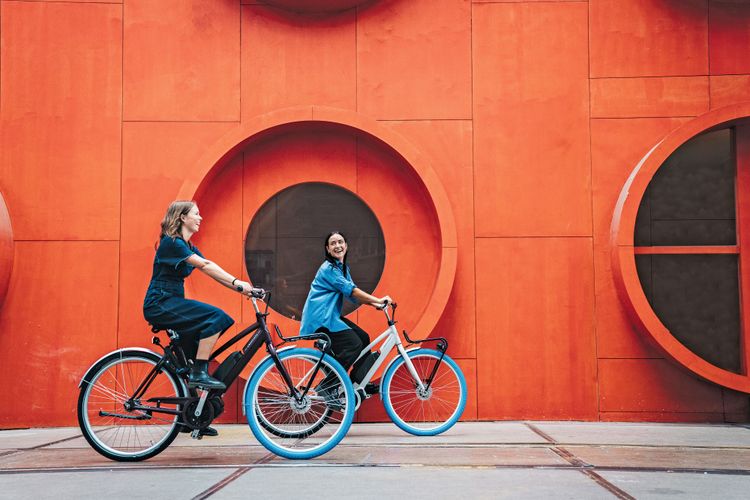 Zwei Frauen auf E-Bikes vor einer orangenen Wand