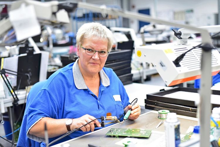 Eine Person in blauer Arbeitskleidung arbeitet an einem Tisch mit elektronischen Komponenten. Sie hält ein Lötwerkzeug und bearbeitet eine Leiterplatte, während sie von hellen Lampen beleuchtet wird. Im Hintergrund ist eine Werkstatt mit weiteren Arbeitsplätzen zu sehen.