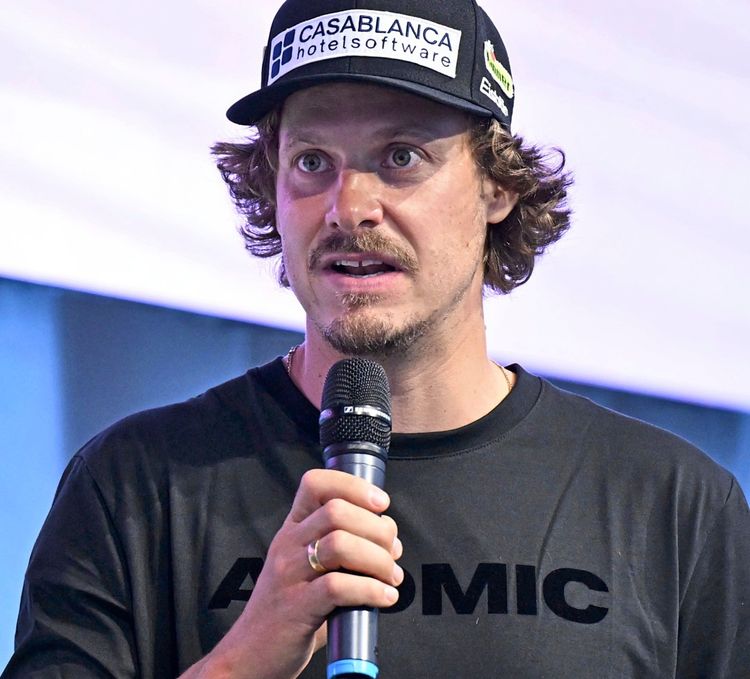 Ein Mann mit schwarzem Basecap und schwarzem Pullover mit der Aufschrift „ATOMIC“ spricht in ein Mikrofon. Hintergrund unscharf.