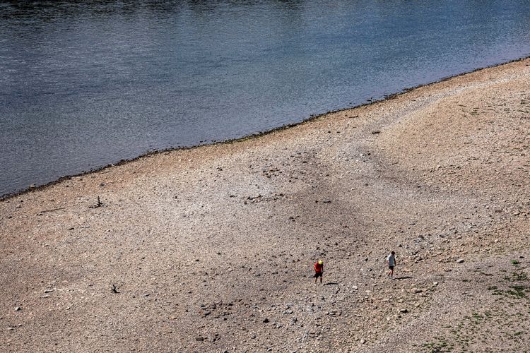 Niedrigwasser am Rhein bei Köln: Blick auf eine trockengelegte Sandbank mit zwei Spaziergängern. Im Hintergrund fließt der stark zurückgegangene Fluss. Die Schifffahrt ist durch den niedrigen Wasserstand eingeschränkt.