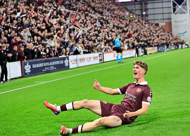 Ein Fußballspieler im dunkelroten Trikot von Heart of Midlothian rutscht jubelnd auf den Knien über den Rasen, nachdem er ein Tor erzielt hat. Im Hintergrund jubeln Fans auf den vollen Tribünen, während ein Schiedsrichter in blau läuft.