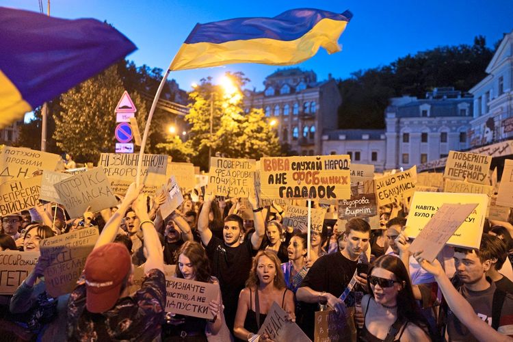 Große Demonstration im öffentlichen Raum während der Abendstunden. Viele Menschen halten Protestschilder mit ukrainischen Aufschriften hoch, während mehrere blau-gelbe ukrainische Flaggen über der Menge wehen. Im Hintergrund sind beleuchtete Gebäude und Straßenlaternen zu sehen.