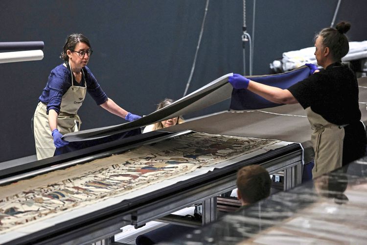 Zwei Museumskuratorinnen, die blaue Handschuhe und beige Schürzen tragen, arbeiten an der Vorbereitung und Enthüllung des mittelalterlichen Bayeux-Teppichs im British Museum in London. Der Teppich, auf einem Tisch ausgerollt, zeigt Szenen der normannischen Eroberung Englands von 1066.