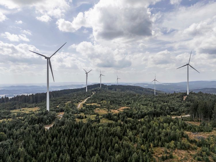 Luftaufnahme eines Windparks mit mehreren Windkraftanlagen auf einem bewaldeten Hügel. Der Himmel ist teils bewölkt, im Hintergrund sind sanfte Hügel der Landschaft in Oberösterreich zu sehen.