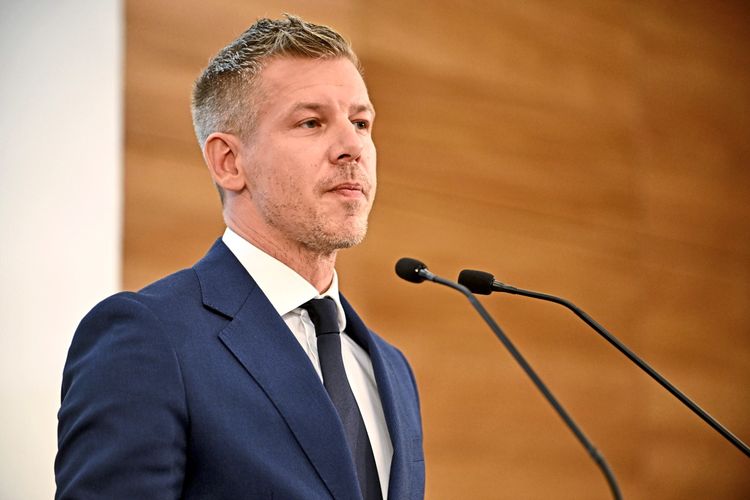 Mann in Anzug und Krawatte steht an einem Rednerpult mit Mikrofonen und spricht auf einer Pressekonferenz. Hintergrund ist verschwommen mit Holzoptik.