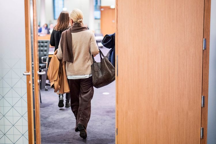 Eine Person geht durch eine geöffnete Tür in einen Raum, in dem eine Sitzung stattfindet. Die Person trägt eine Handtasche und eine Jacke über dem Arm. Im Hintergrund sitzen Menschen an einem Tisch.