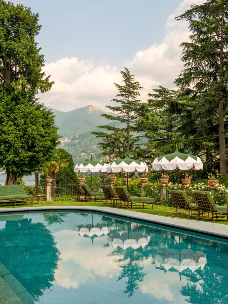 Swimmingpool eines Hotels mit Grünanlage und Sonneschirmen und Sonnenliegen, im Hintergrund Berge