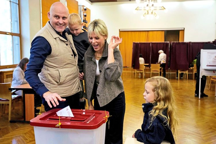Eine Familie gibt ihre Stimmen während einer Wahl in einem Wahllokal ab. Ein Mann steckt einen Wahlzettel in eine Wahlurne, während eine Frau und zwei Kinder danebenstehen. Der Raum ist mit Holzverkleidungen und Wahlkabinen im Hintergrund ausgestattet.