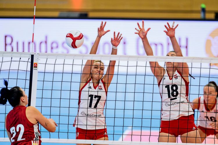 Eine Volleyball-Testspielszene der Frauen zwischen Österreich und Deutschland am 15. August 2026 in Wien. Zwei österreichische Spielerinnen (Nr. 17 und Nr. 18) blockieren am Netz, während eine deutsche Spielerin (Nr. 22) angreift. Im Hintergrund ist das Spielfeld und ein unscharfes Werbebanner zu sehen.