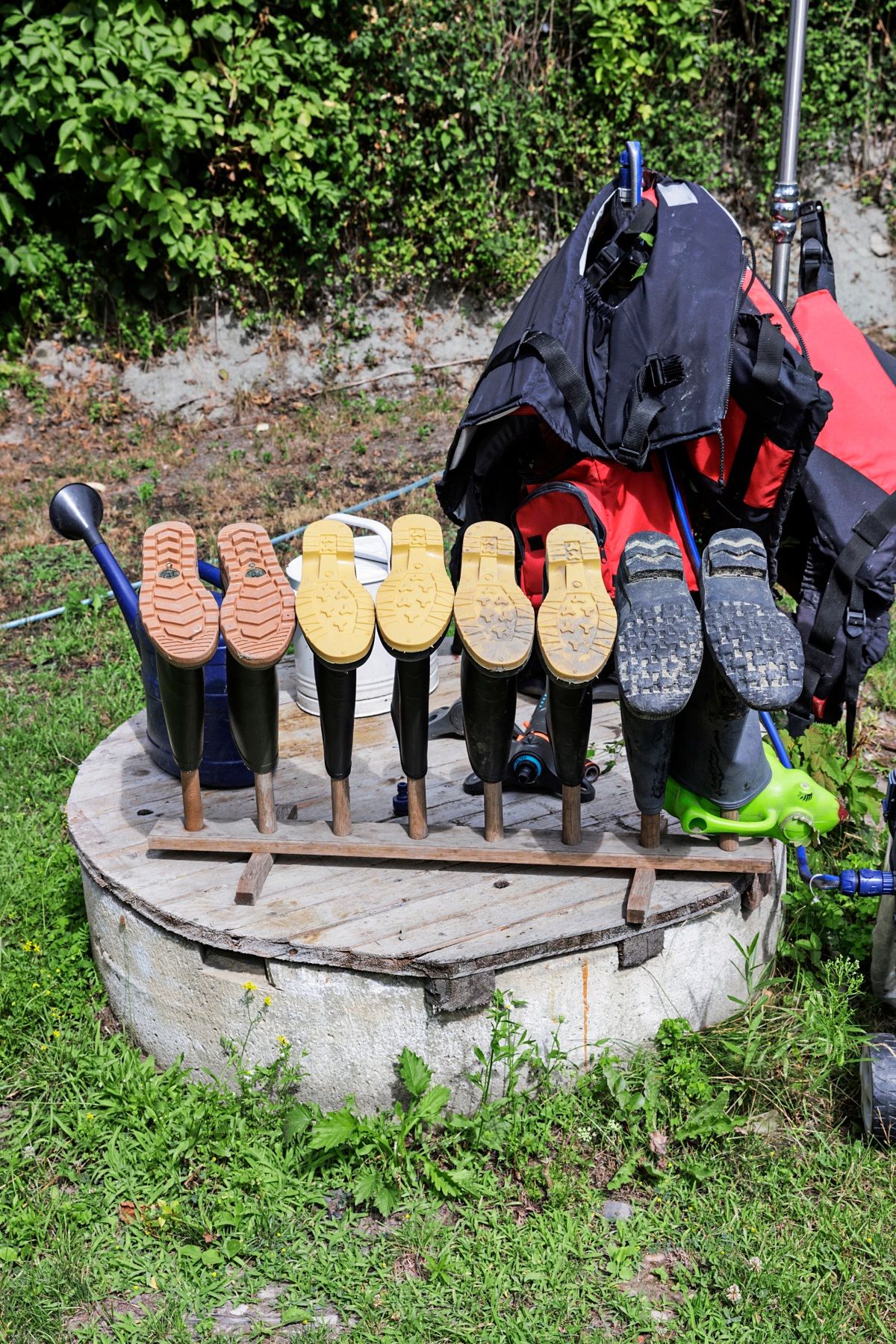 Auf dem Bild sind mehrere Gummistiefel zu sehen, die umgedreht auf einem Holzgestell zum Trocknen aufgestellt sind. Im Hintergrund befinden sich grüne Büsche und Gras, und daneben liegen rote und schwarze Ausrüstungsgegenstände.