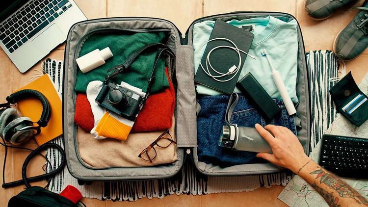 Ein Mann packt einen Koffer, der offen auf dem Boden liegt und mit Kleidung, Zahnbürste und elektronischen Geräten gefüllt ist