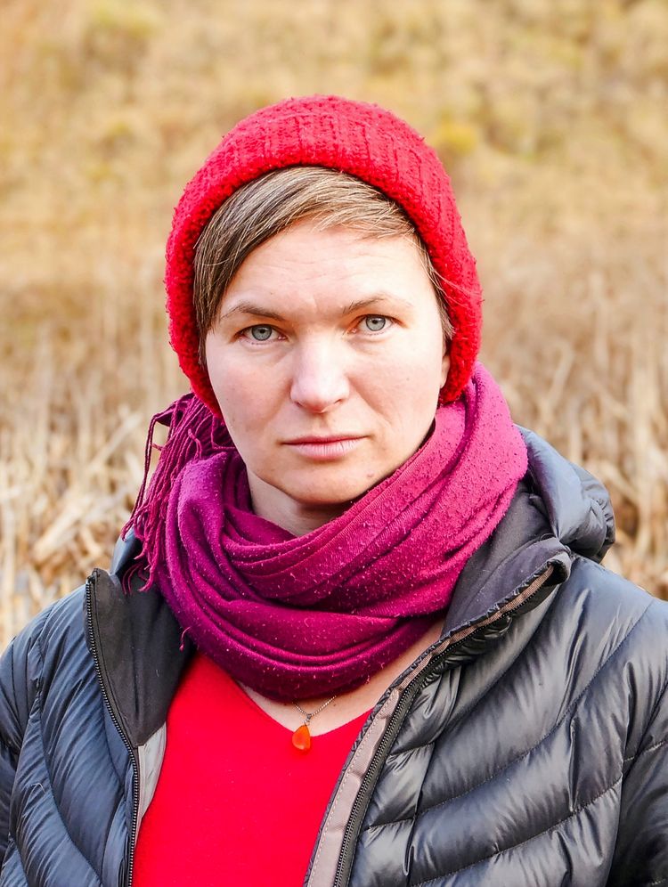 Eine Person mit roter Strickmütze, bordeauxfarbenem Schal und schwarzer Steppjacke blickt direkt in die Kamera. Der Hintergrund ist unscharf und zeigt eine herbstliche Landschaft.