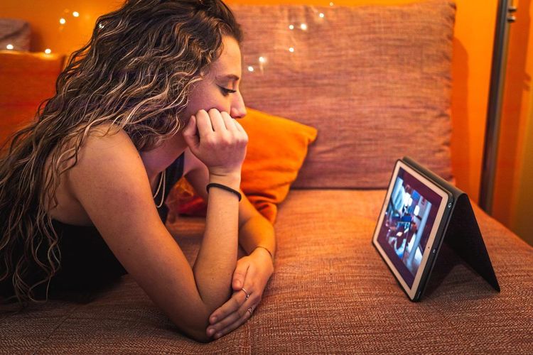 Eine junge Frau schaut bäuchlings auf einem Sofa liegend auf einem Tablet Serien an