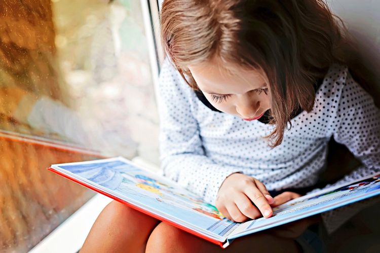 Ein kleines Mädchen sitzt in der Nähe eines Fensters und liest ein buntes Buch, während ihr Finger auf einer Seite zeigt.