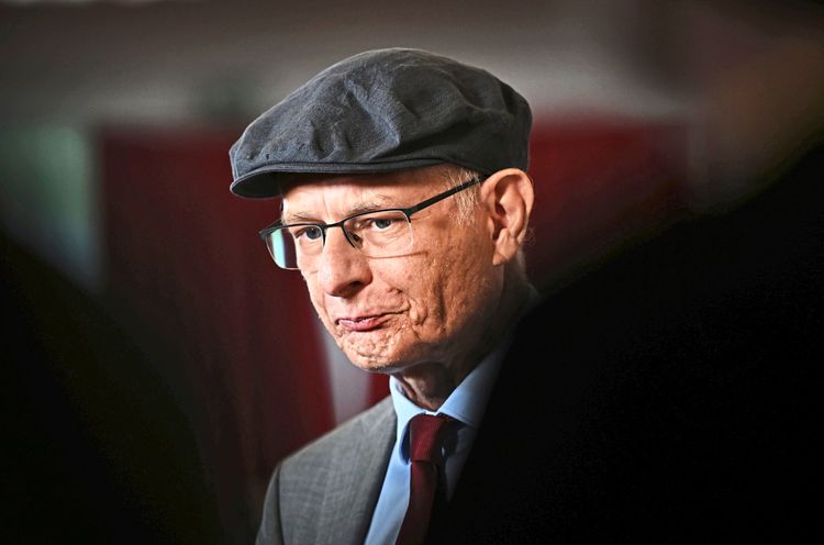 Finanzminister Markus Marterbauer mit Brille und dunkler Schiebermütze, gekleidet in einen Anzug mit roter Krawatte, blickt nachdenklich. Der Hintergrund ist unscharf.