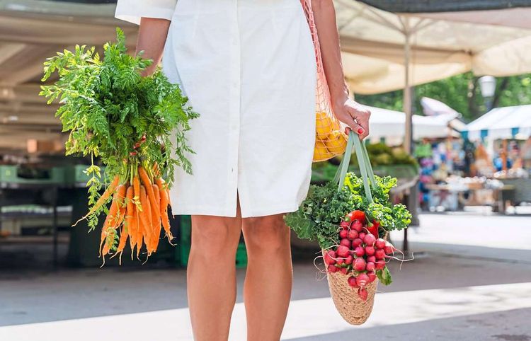 Eine Frau trägt Taschen mit Nahrungsmitteln, im Hintergrund sind Marktstände zu sehen.