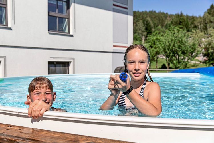 Zwei Kinder spielen in einem Aufstellpool im Freien. Eines der Kinder hält eine Wasserpistole. Im Hintergrund sind ein Gebäude, Bäume und eine Wiese zu sehen.