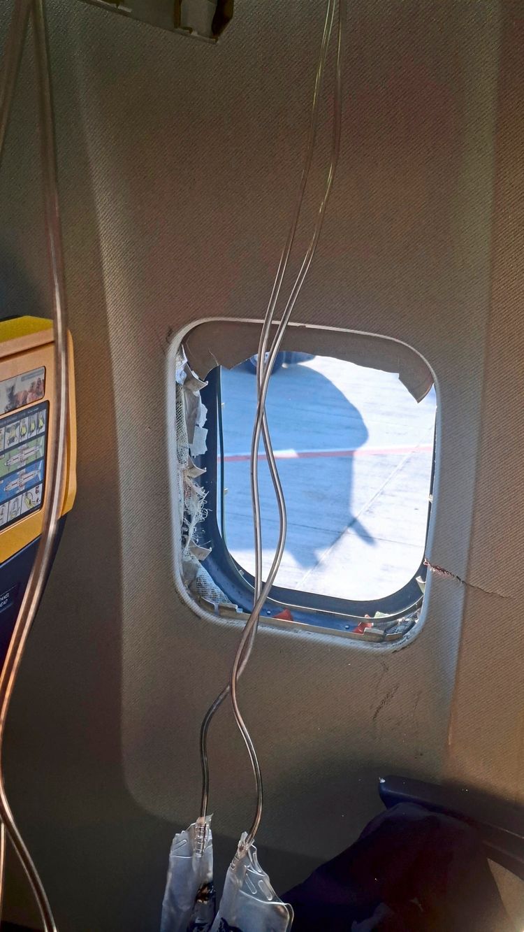 Ein beschädigtes Fenster in der Kabine eines Flugzeugs, vermutlich einer Ryanair-Maschine, mit freigelegten Kabeln. Das Fenster zeigt deutliche Schäden am Rahmen. Im Hintergrund ist der Boden des Flugplatzes sichtbar.