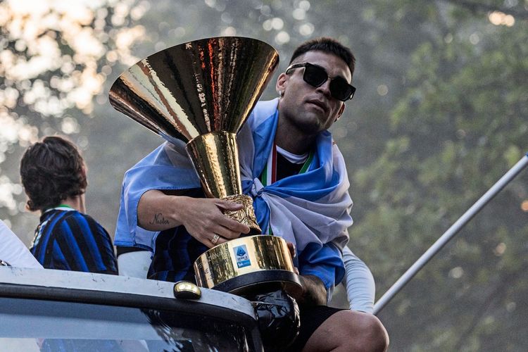 Ein Fußballspieler in einem blauen und schwarzen Trikot sitzt auf einem offenen Bus und hält eine große, goldene Trophäe. Er trägt eine Sonnenbrille und hat eine Flagge über seine Schultern gelegt. Im Hintergrund sind weitere Personen und Bäume zu sehen. Die Szene zeigt eine Meisterfeier.