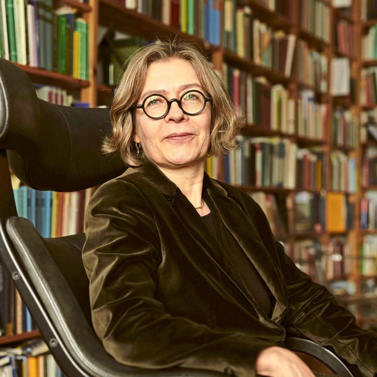 Eine Frau mit Brille, kurzem blondem Haar und einem dunkelgrünen Samtblazer sitzt auf einem schwarzen Stuhl vor einer Bücherwand.