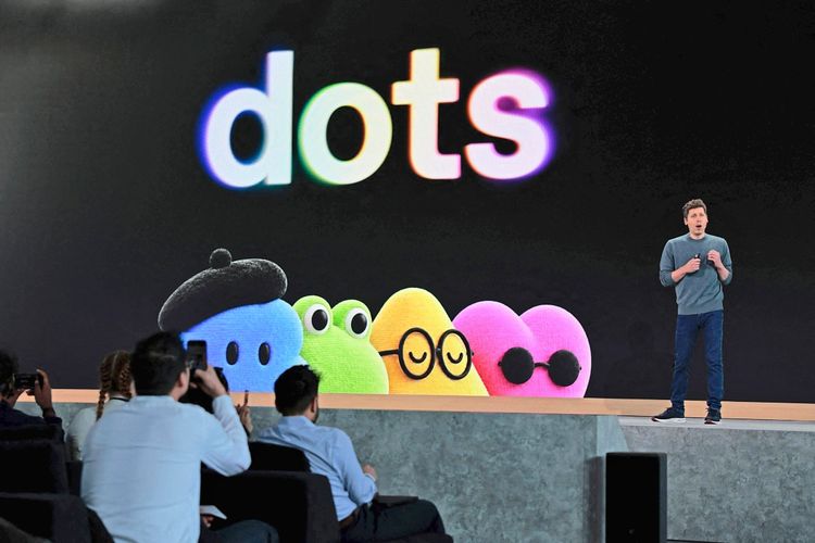 Ein Redner steht auf einer Bühne während einer Präsentation. Im Hintergrund ist ein großes Display mit dem Wort „dots“ sowie bunten, cartoonartigen Figuren in verschiedenen Farben und Formen zu sehen. Im Vordergrund sitzen Zuschauer, einige machen Fotos.