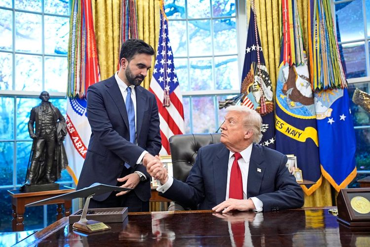 Zwei Männer schütteln sich im Oval Office des Weißen Hauses die Hände. Einer der Männer sitzt hinter dem Schreibtisch, während der andere steht. Im Hintergrund sind amerikanische Flaggen, militärische Wimpel und eine Statue zu sehen.