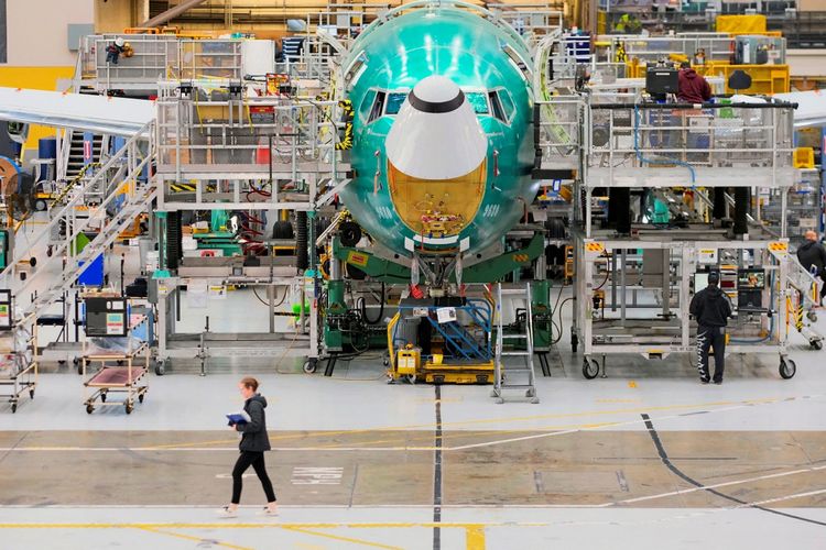 Eine teilweise montierte Boeing 737 MAX befindet sich auf der Endmontagelinie in einer Fabrikhalle. Mitarbeiter arbeiten an der Maschine, während eine Person im Vordergrund entlanggeht.