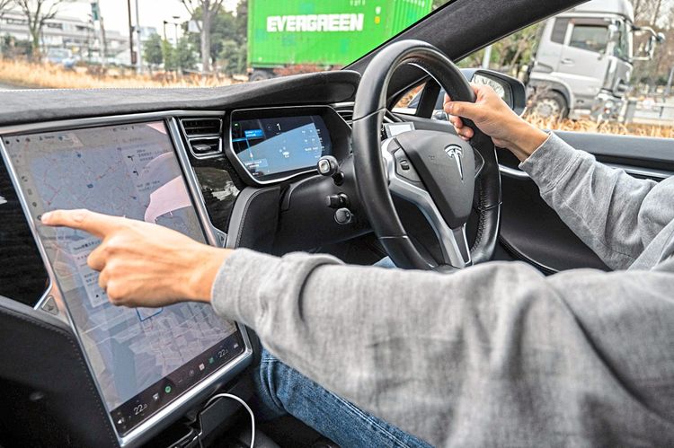 Eine Innenraumaufnahme einse Tesla Model S mit Lenkrad auf der rechten Seiten. Der Fahrer greift auf den Touchscreen, wo eine Karte der Umgebung dargestellt wird. Er trägt einen grauen Pullover und eine blaue Jeans. Es handelt sich um Atsushi Ikeda, den Vizepräsidenten des Tesla-Clubs in Japan. Er ist in Tokio unterwegs, im Hintergrund ist ein Laster mit einem Evergreen-Schriftzug zu sehen.