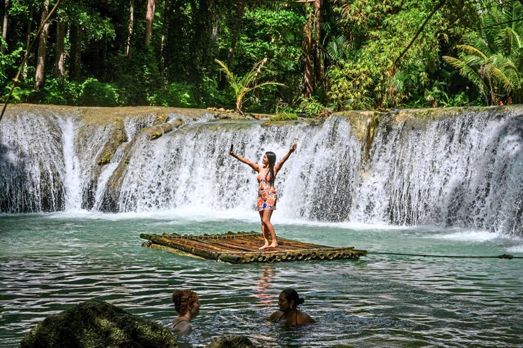 Eine Frau steht auf einem Bambusfloß vor den Cambugahay-Wasserfällen auf der Insel Siquijor, Philippinen, und macht ein Selfie. Im Wasser schwimmen zwei weitere Personen. Die Szenerie ist von üppiger tropischer Vegetation umgeben.