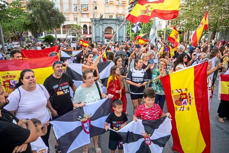 Eine Menschenmenge versammelt sich bei einer Demonstration in Ceuta, Spanien. Viele tragen spanische Flaggen oder Flaggen von Ceuta und protestieren gegen die Immigrationssituation. Die Szene spielt sich auf einem öffentlichen Platz ab, umgeben von Bäumen und Gebäuden.
