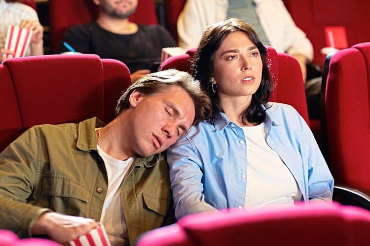 Ein Mann und eine Frau sitzen im Kino, sie schaut mit starrem Blick auf die Leinwand, während er an ihrer Schulter eingeschlafen ist, die Hand im Popcornstanitzel